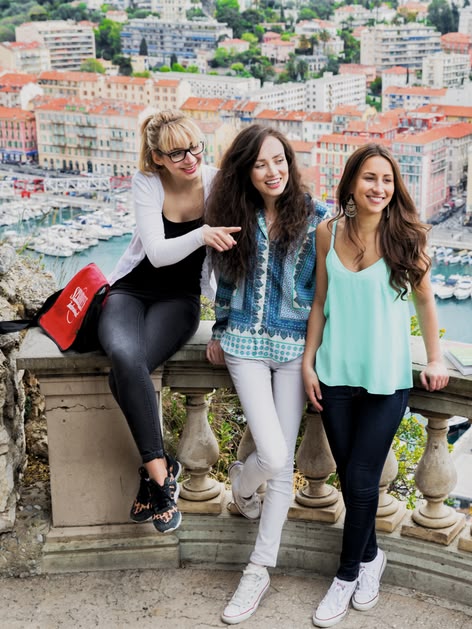 Drei Sprachcaffe-Schülerinnen lächeln auf einem Balkon mit Blick auf Nizza, Frankreich, mit dem Hafen und den bunten Gebäuden ringsum