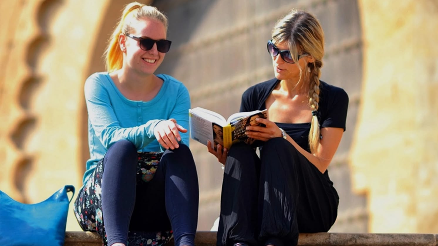 Zwei Frauen sitzen auf Steinstufen und lesen ein Buch in Rabat, Marokko, mit historischer Architektur hinter ihnen.