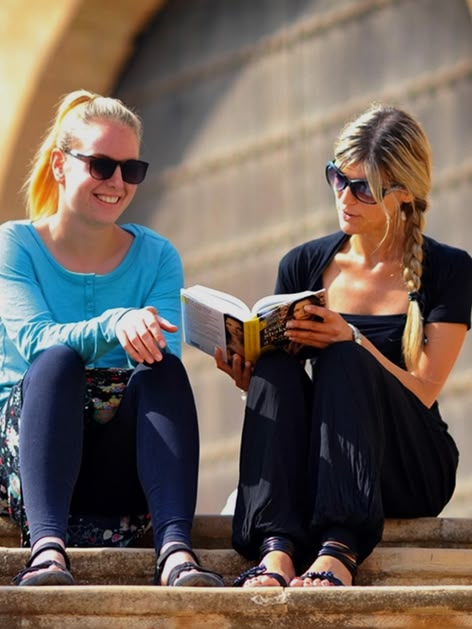 Two women sitting on stone steps reading a book in Rabat, Morocco, with historic architecture behind them.