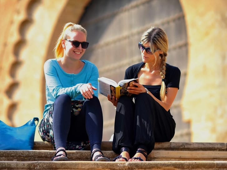 Two women sitting on stone steps reading a book in Rabat, Morocco, with historic architecture behind them.