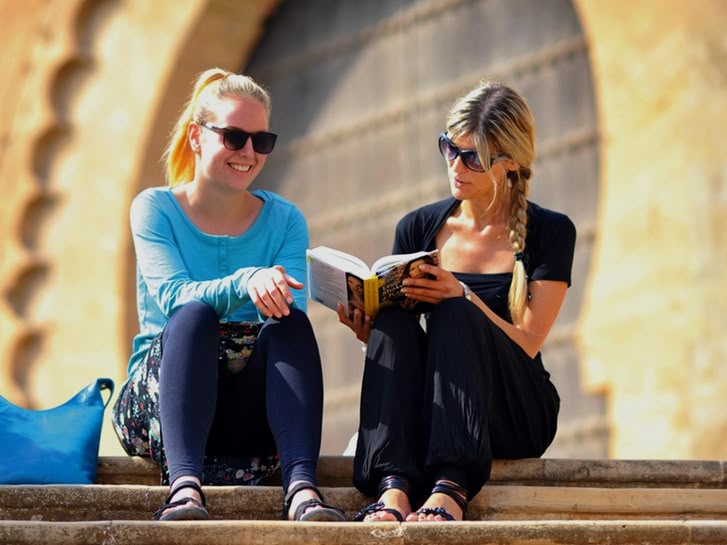 Two Sprachcaffe students sitting on steps in Rabat, Morocco, reading and chatting in front of traditional architecture.