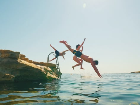 Eine Gruppe Jugendlicher springt an einem sonnigen Tag von den Felsen ins Meer.