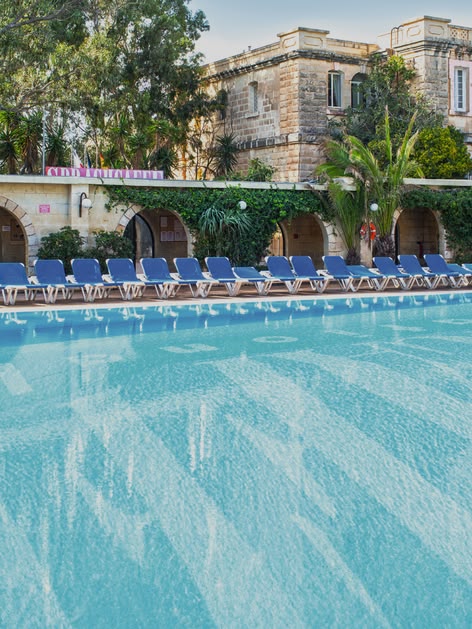 Large outdoor swimming pool surrounded by sun loungers and Mediterranean-style buildings at Sprachcaffe Malta.