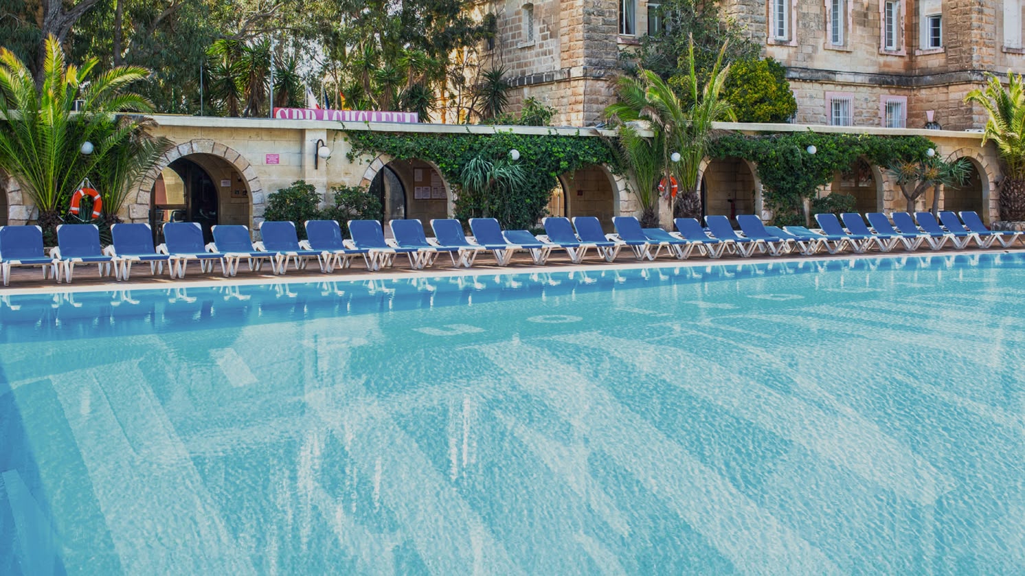 Large outdoor swimming pool surrounded by sun loungers and Mediterranean-style buildings at Sprachcaffe Malta.