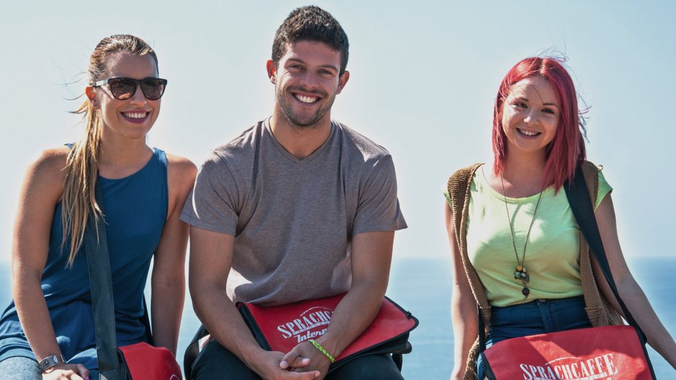 Drei Schüler mit Sprachcaffe-Tüten sitzen auf einem Geländer und lächeln mit dem Meer im Hintergrund in Málaga, Spanien.