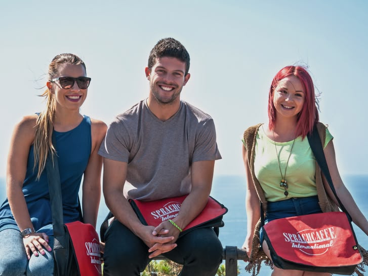 Three students with Sprachcaffe bags sitting on a railing, smiling with the sea in the background in Málaga, Spain.