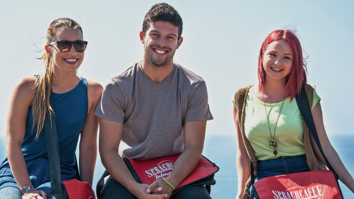 Drei Schüler mit Sprachcaffe-Tüten sitzen auf einem Geländer und lächeln mit dem Meer im Hintergrund in Málaga, Spanien.
