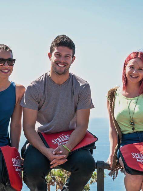 Three students with Sprachcaffe bags sitting on a railing, smiling with the sea in the background in Málaga, Spain.