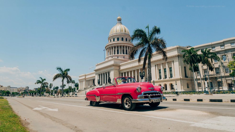 Ein klassisches rosafarbenes Auto fährt an El Capitolio in Havanna, Kuba, mit Palmen und blauem Himmel vorbei.