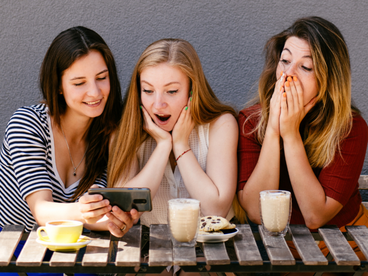 Eine Gruppe von Frauen schauen sich mit erstaunten und frohen Ausdrücken die erklärung zum Thema Simple Past oder Present Perfect an. Sie sitzen mit Milchkaffe an einem Tisch. Der Hintergrund ist eine graue Wand. 