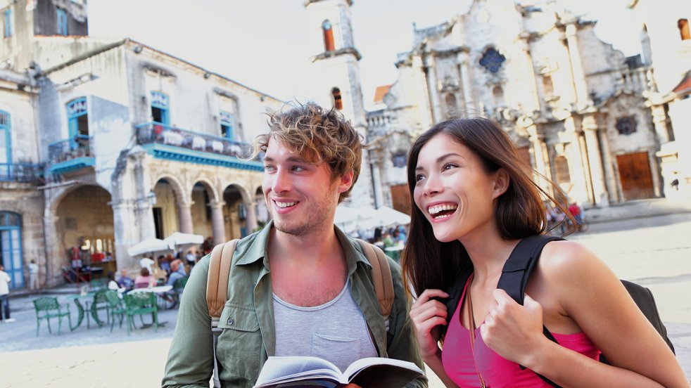 Zwei Touristen, die Alt-Havanna, Kuba, erkunden, halten einen Reiseführer in der Hand und lächeln auf einem historischen Platz mit kolonialer Architektur.