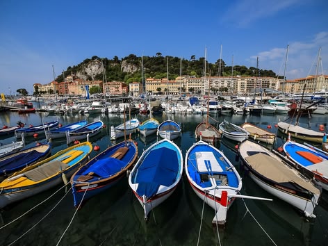 Bunte Boote im Hafen von Nizza, Frankreich, mit Gebäuden am Hang und einem Jachthafen im Hintergrund.