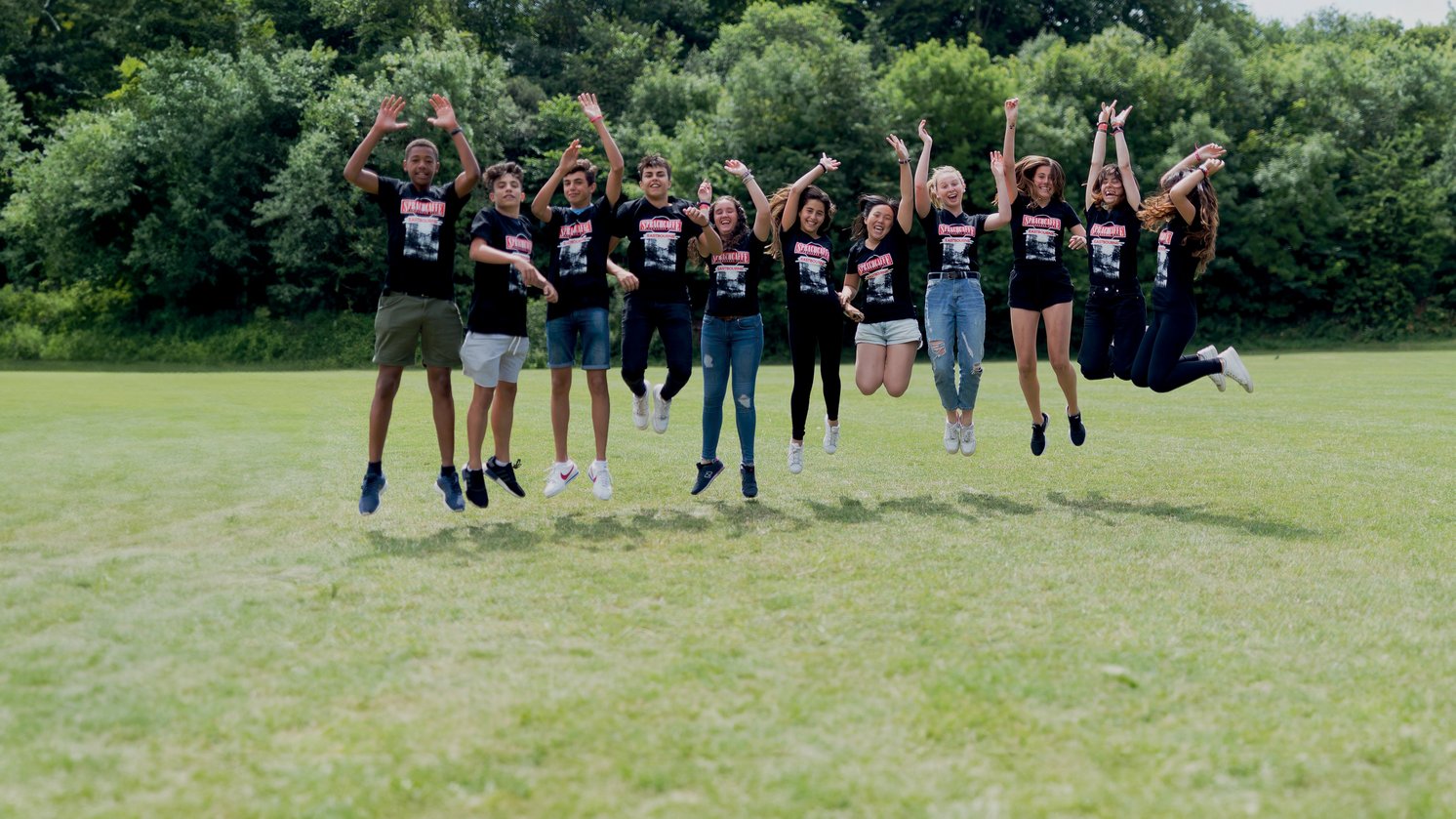 Sprachcaffe-Schülerinnen und -Schüler in England springen in der Luft auf einer grünen Wiese, tragen passende Shirts und genießen die freie Natur.