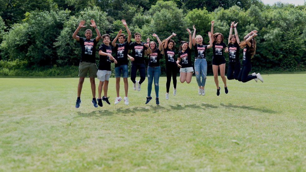 Sprachcaffe-Schülerinnen und -Schüler in England springen in der Luft auf einer grünen Wiese, tragen passende Shirts und genießen die freie Natur.