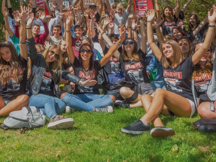 Group of Sprachcaffe students sitting on the grass in London, smiling and raising their arms during an outdoor activity.