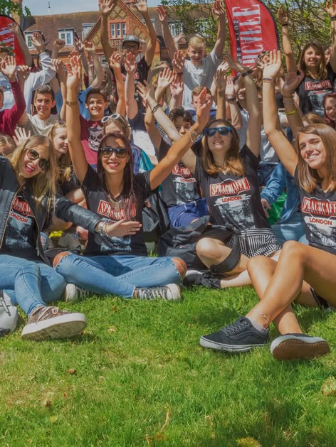 Group of Sprachcaffe students sitting on the grass in London, smiling and raising their arms during an outdoor activity.