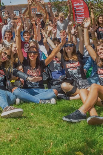 Eine Gruppe von Sprachcaffe-Schülerinnen und -Schülern sitzt auf dem Rasen in London, lächelt und hebt die Arme während einer Outdoor-Aktivität.