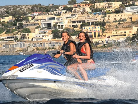 Zwei junge Frauen lächeln, während sie mit einem Jetski an der Küste Maltas entlangfahren.