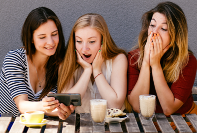 Eine Gruppe von Frauen schauen sich mit erstaunten und frohen Ausdrücken die erklärung zum Thema Simple Past oder Present Perfect an. Sie sitzen mit Milchkaffe an einem Tisch. Der Hintergrund ist eine graue Wand. 