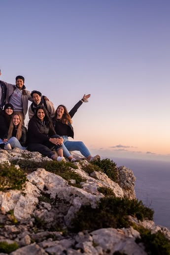 Group of young adults on a rocky cliff in Malta at sunset, overlooking the sea and enjoying the view.