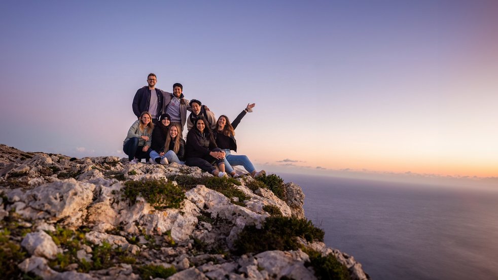 Gruppe junger Erwachsener auf einer felsigen Klippe in Malta bei Sonnenuntergang, die das Meer überblickt und die Aussicht genießt.