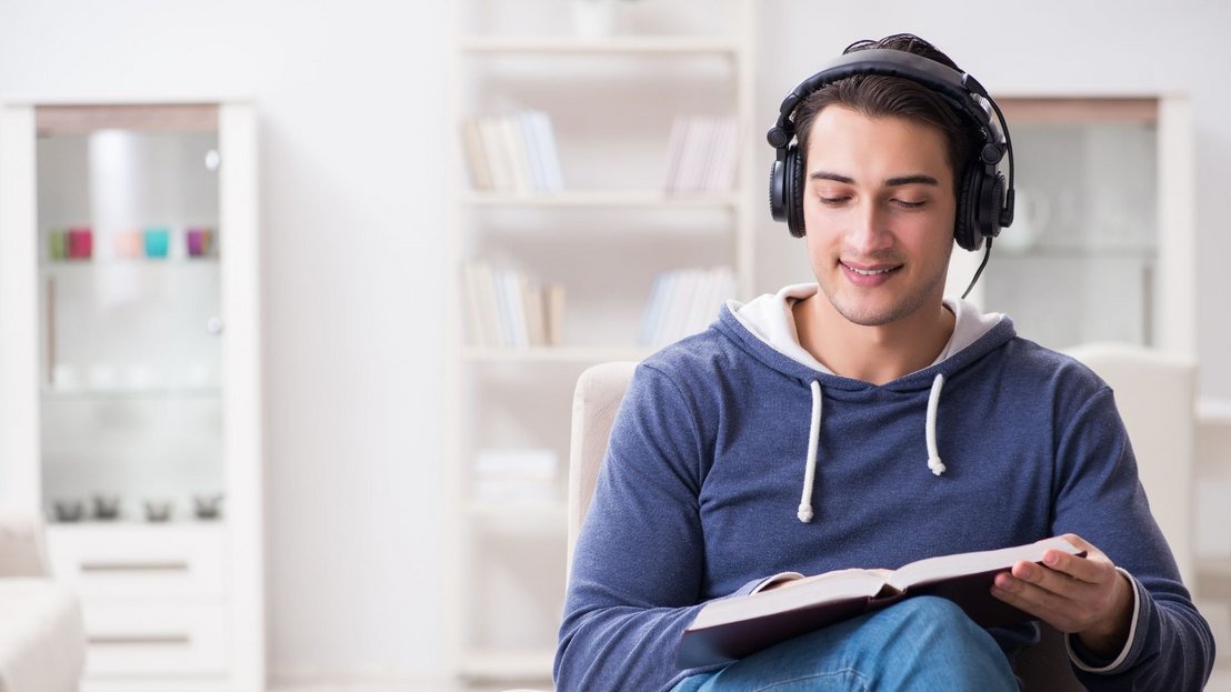 Hier sieht einen jungen Mann im blauen Pullover. Er lernt englisch indem er sowohl liest als auch mit Kopfhörern dem Text zuhört.