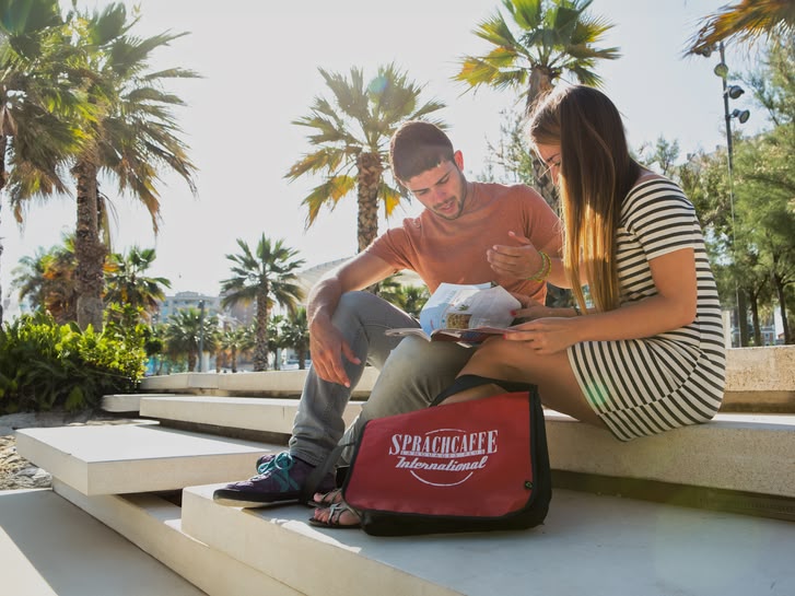 Two students sitting by the seaside in Spain with a Sprachcaffe bag, surrounded by palm trees and modern steps.